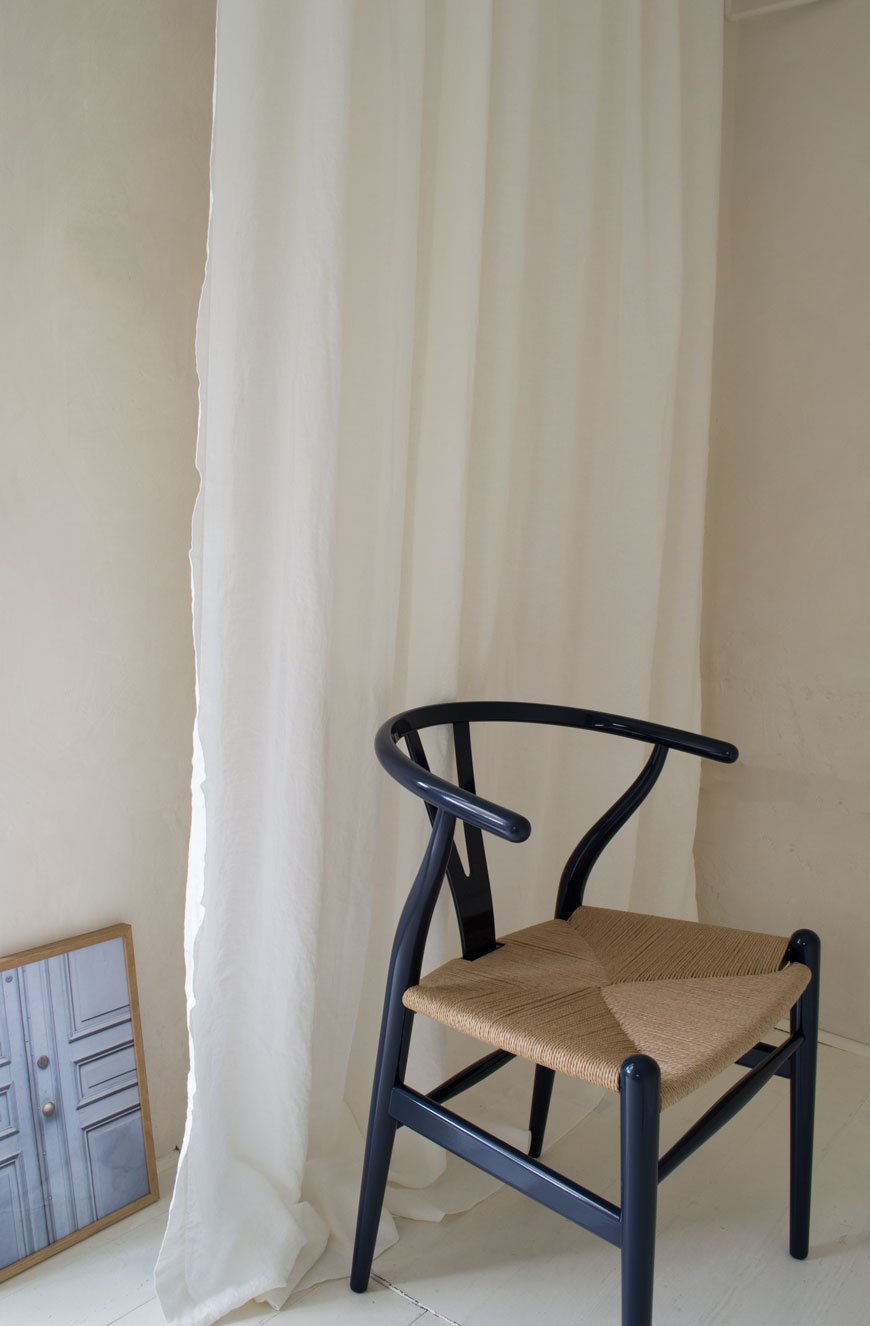 A minimalist scene with a navy blue Hans J.Wegner chair in front of off-white linen curtains and a framed image of weathered blue doors. 