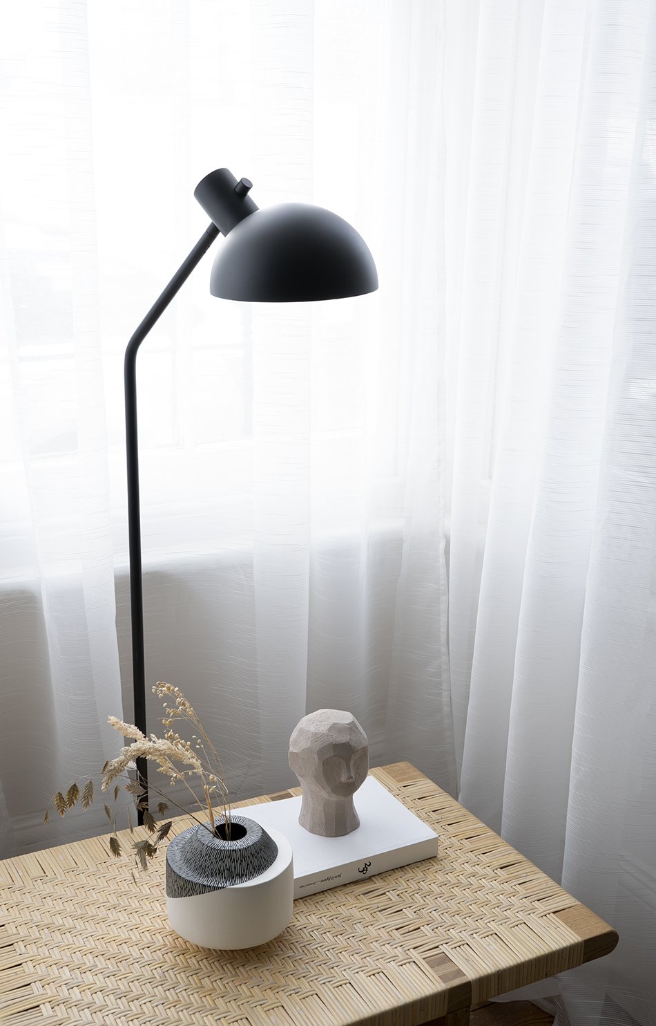Ceramic objects styled on a rattan bench in a bay window with a minimalist black floor lamp behind. 