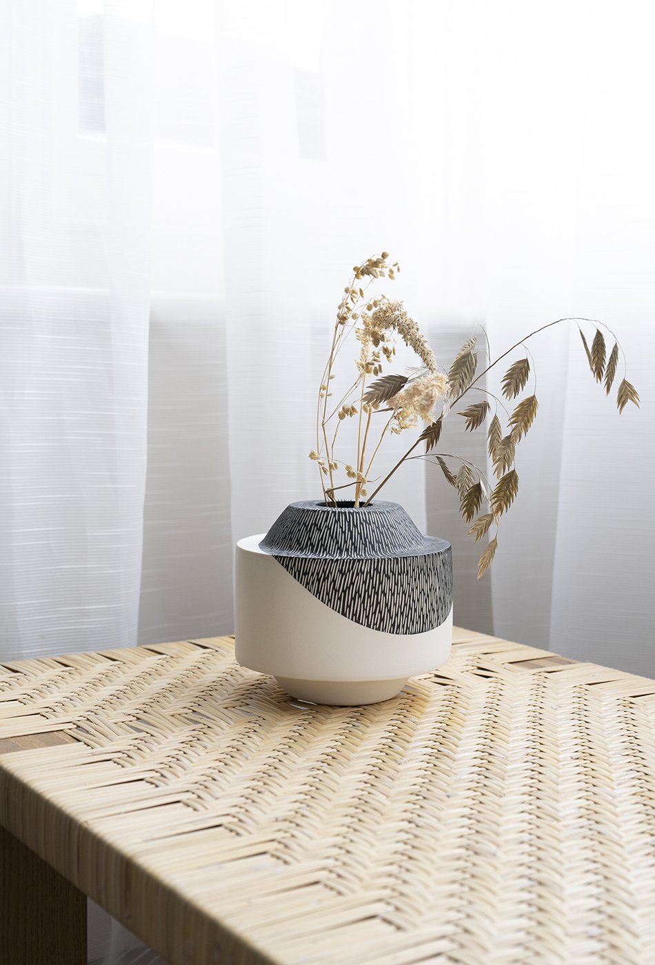 A carved monochrome black and white ceramic vase made by British ceramist Sophie Bland sits on top of a woven rattan table bench designed by Børge Mogensen. 