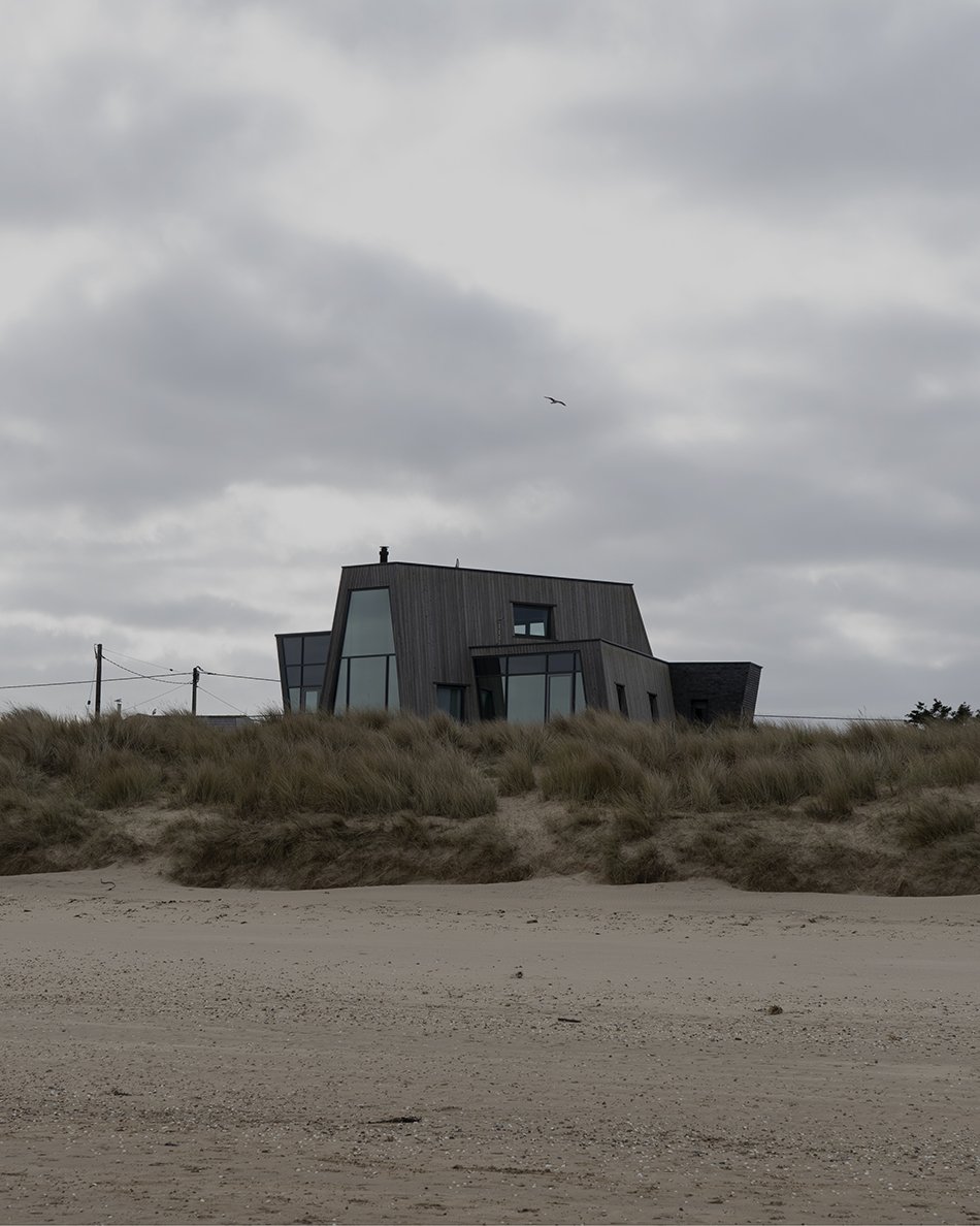 A contemporary beach house clad in larch wood with large windows on the grassy dunes of sandy beach at Greatstone, Kent. 