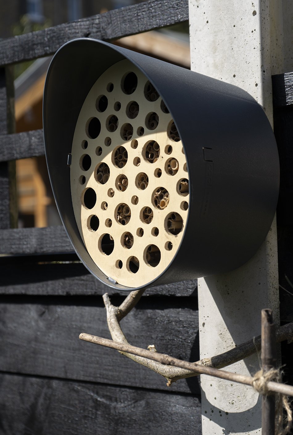 A contemporary oval shape bee hotel designed by Capi for solitary bees, hung on a concrete fence post in a garden. 