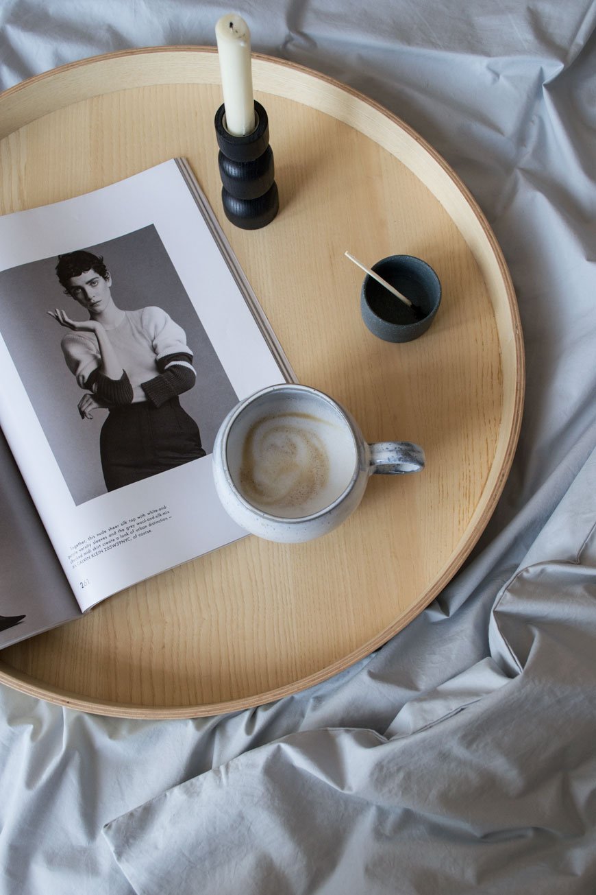 round wooden tray with a cup of coffee and a magazine on cotton sheets on the bed