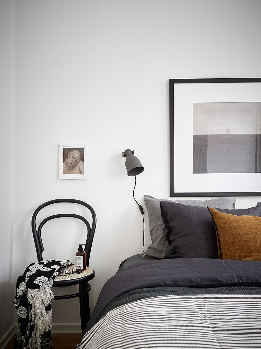 Black framed art and black wall lamp in a minimalist white bedroom with a black bentwood chair for a bedside table.