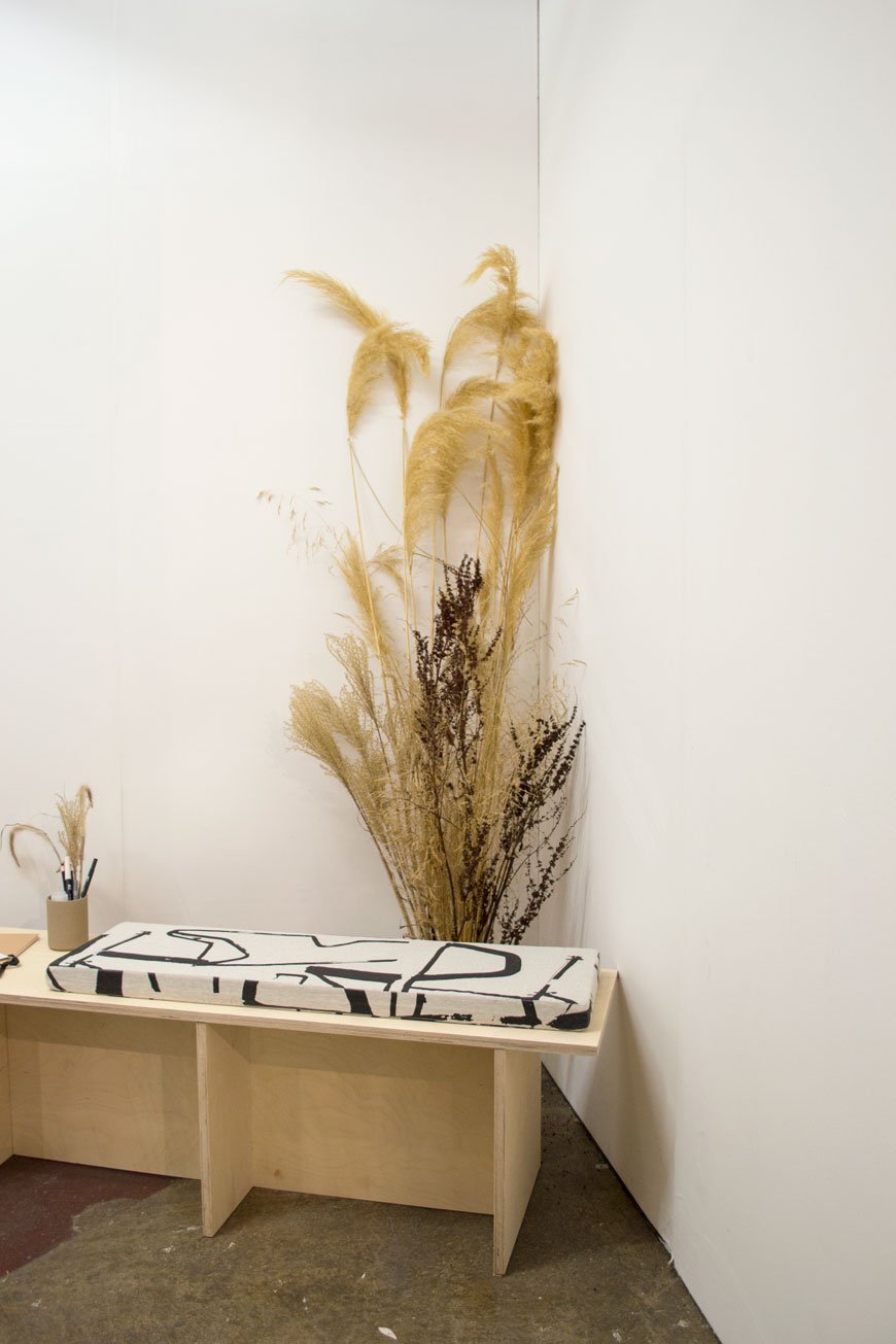 A vase of tall grasses stand behind a plywood bench at Laura Slater's LAND collaboration at LDF