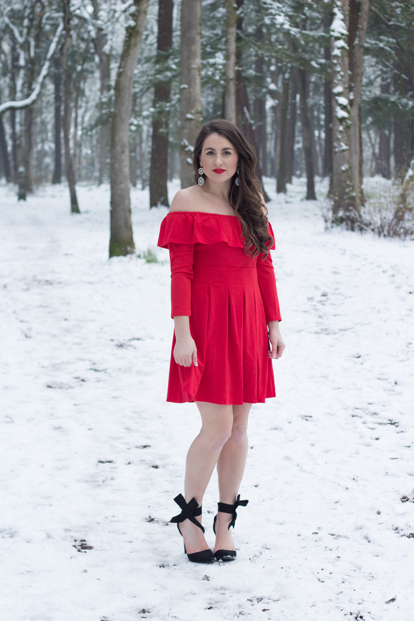 A classic Winter look in a black pea coat, bow heels, and a red off the shoulder dress in the snow.