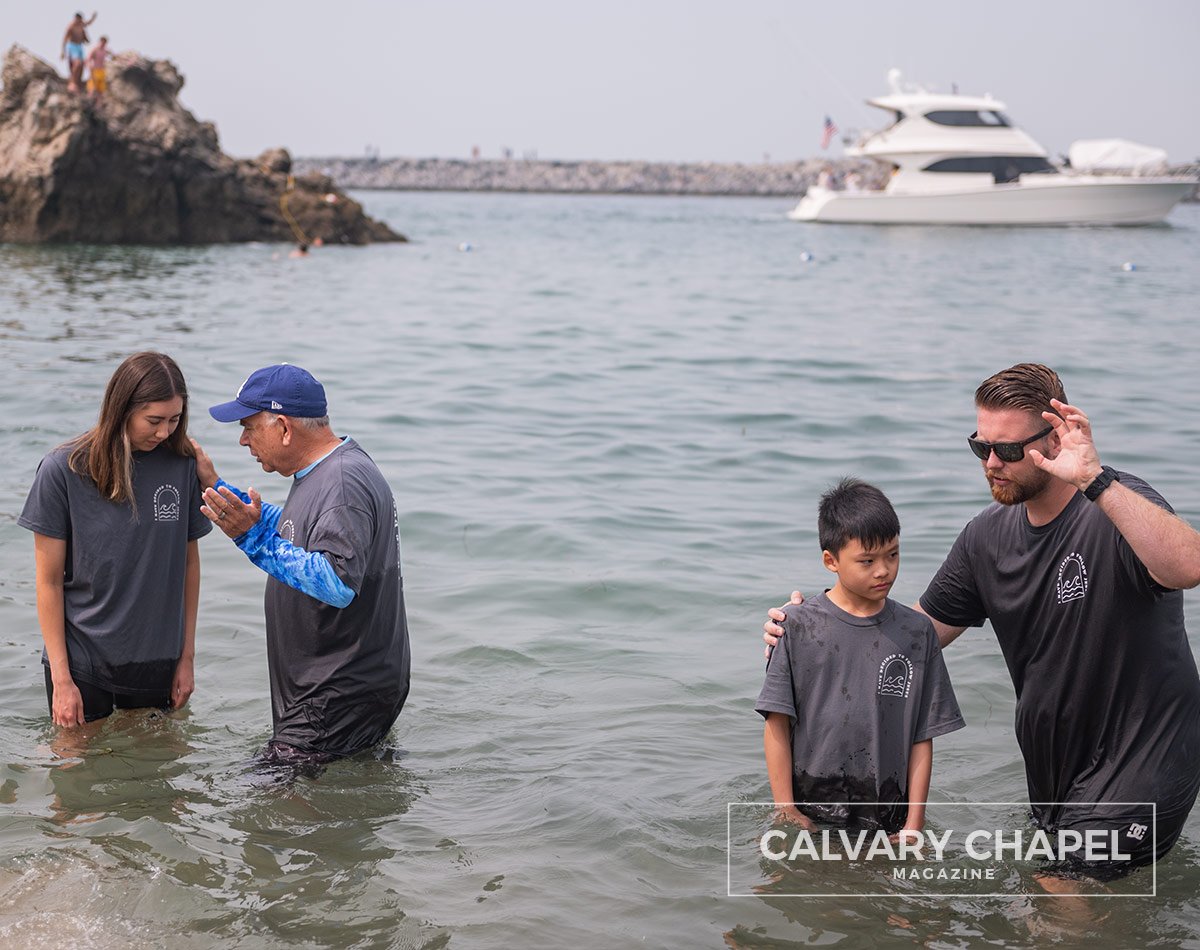 Men baptize kids with boat in background and kids jumping off rocks