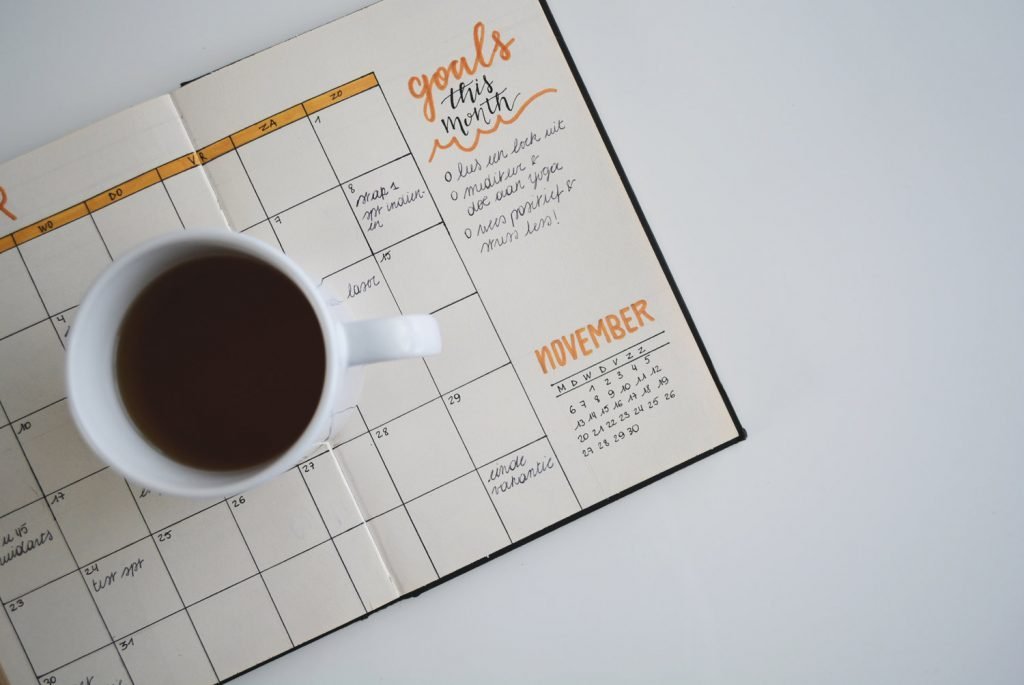 White background with planner opened to a calendar and a coffee mug sits on top of the open planner.