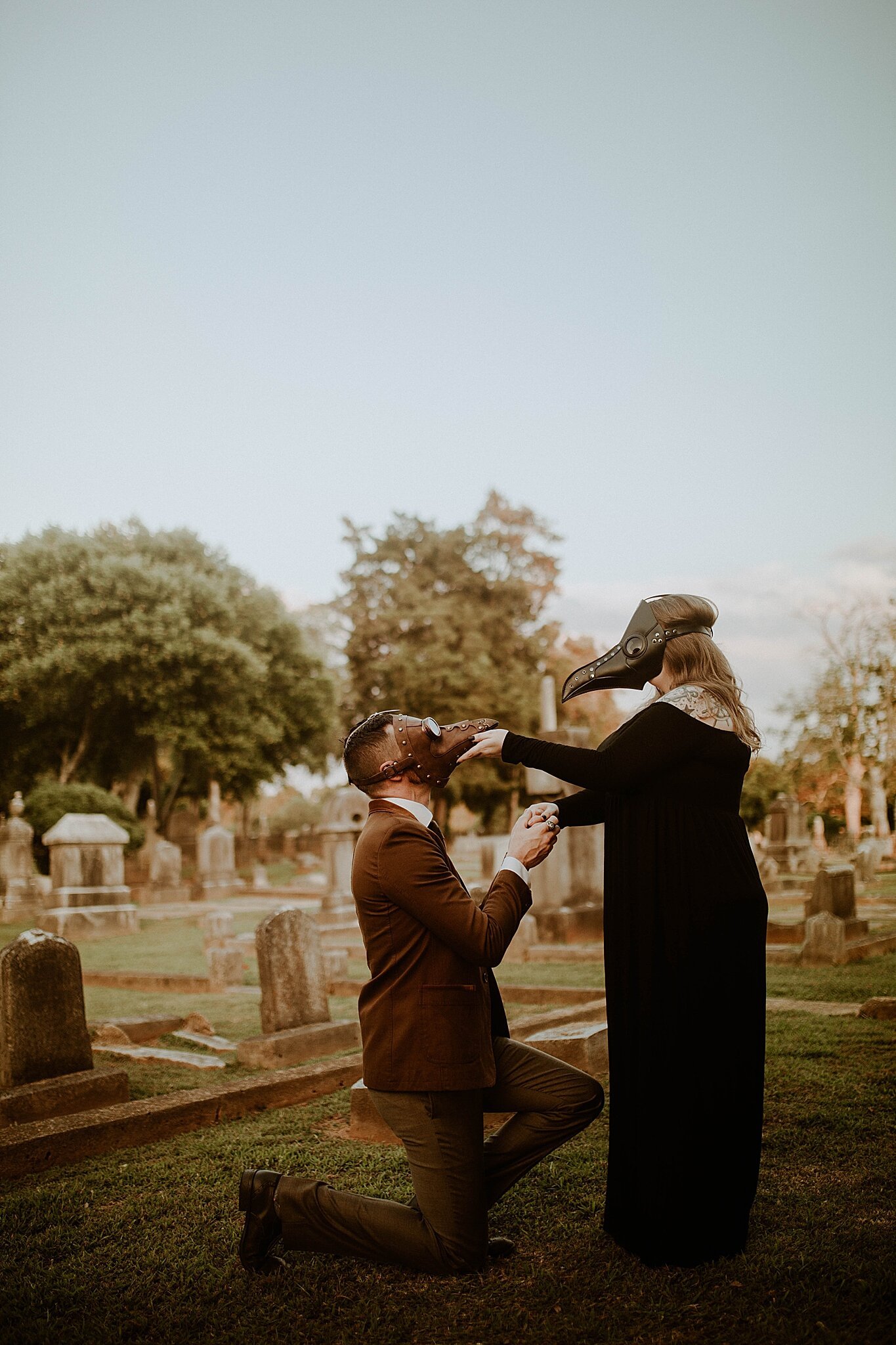 groom in plague mask kneels before fiancee during graveyard engagement session