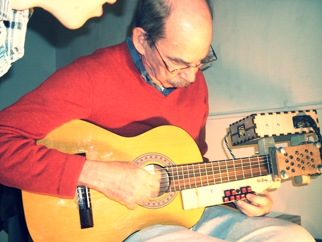 Photo of a man playing a modified guitar with buttons on the fretboard while another man looks on. 