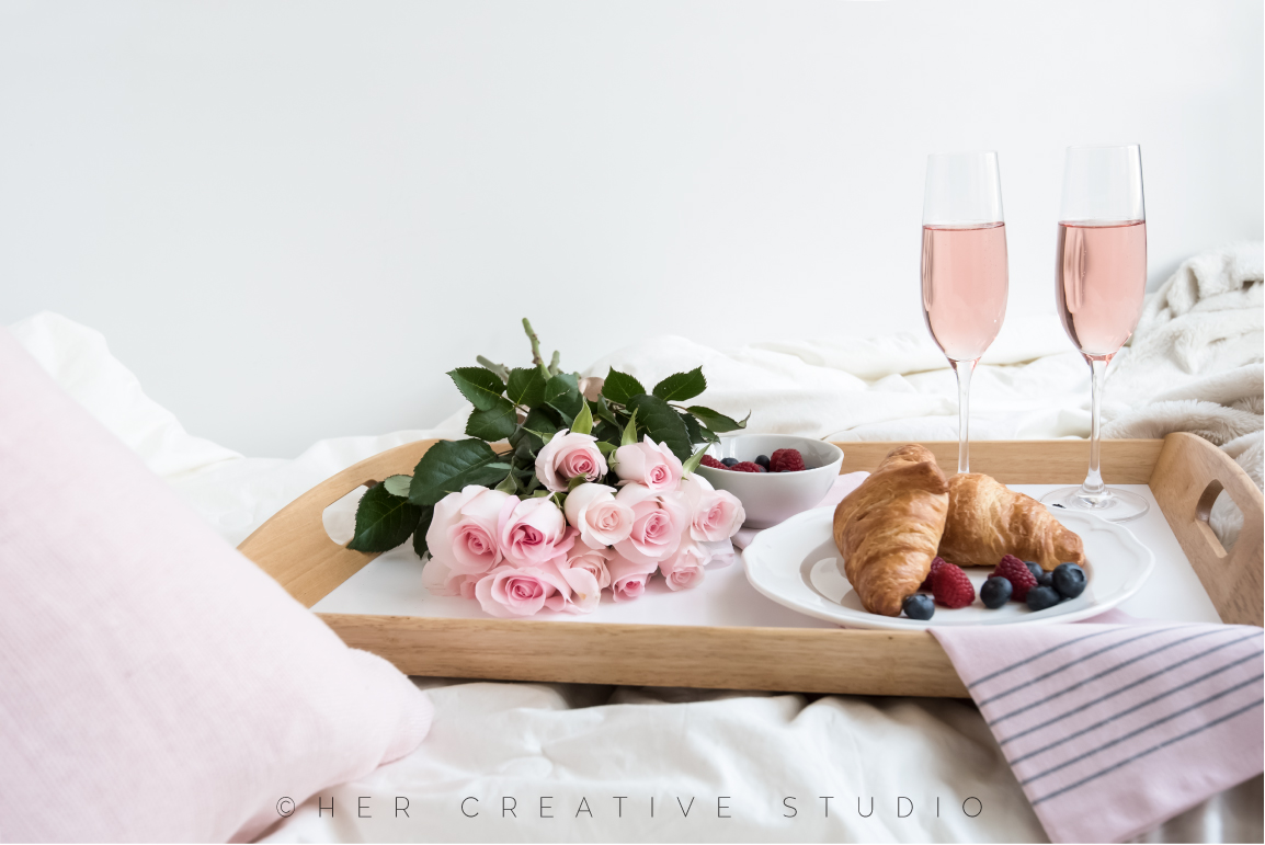 A tray with croissants, berries, pink roses and two glasses of pink champagne sitting on a bed. 
Breakfast-in-bed-1-IE.jpg