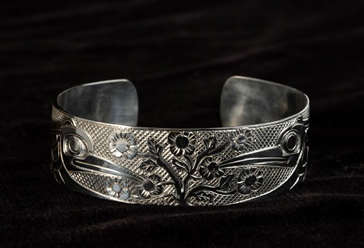 This 3/4 inch wide by 6 1/2 inches long bracelet was created by Haida artist Carmen Goertzen. The design is two hummingbirds feasting on local salmonberry flowers.