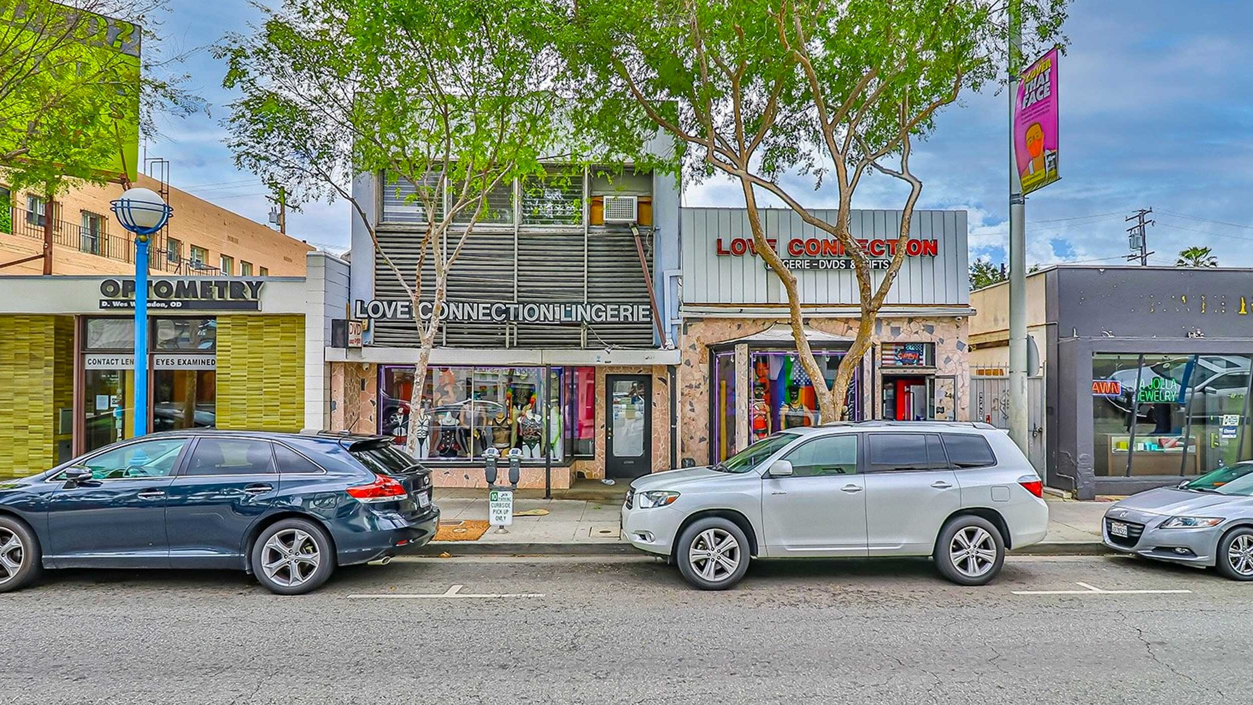 Vibrant storefronts with parked cars and green trees on a city street.