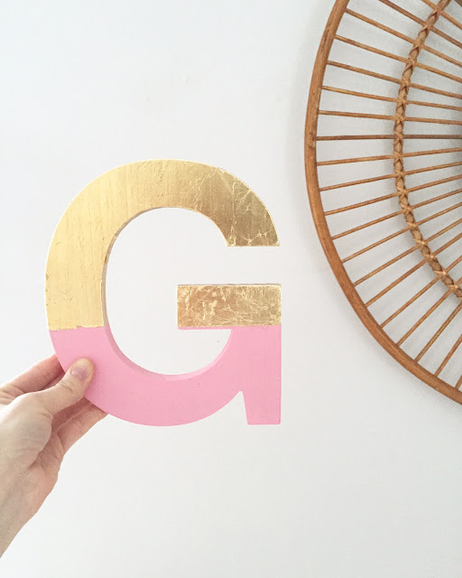 A hand holds up a wooden letter G that's half painted light pink and half covered in golf leaf.