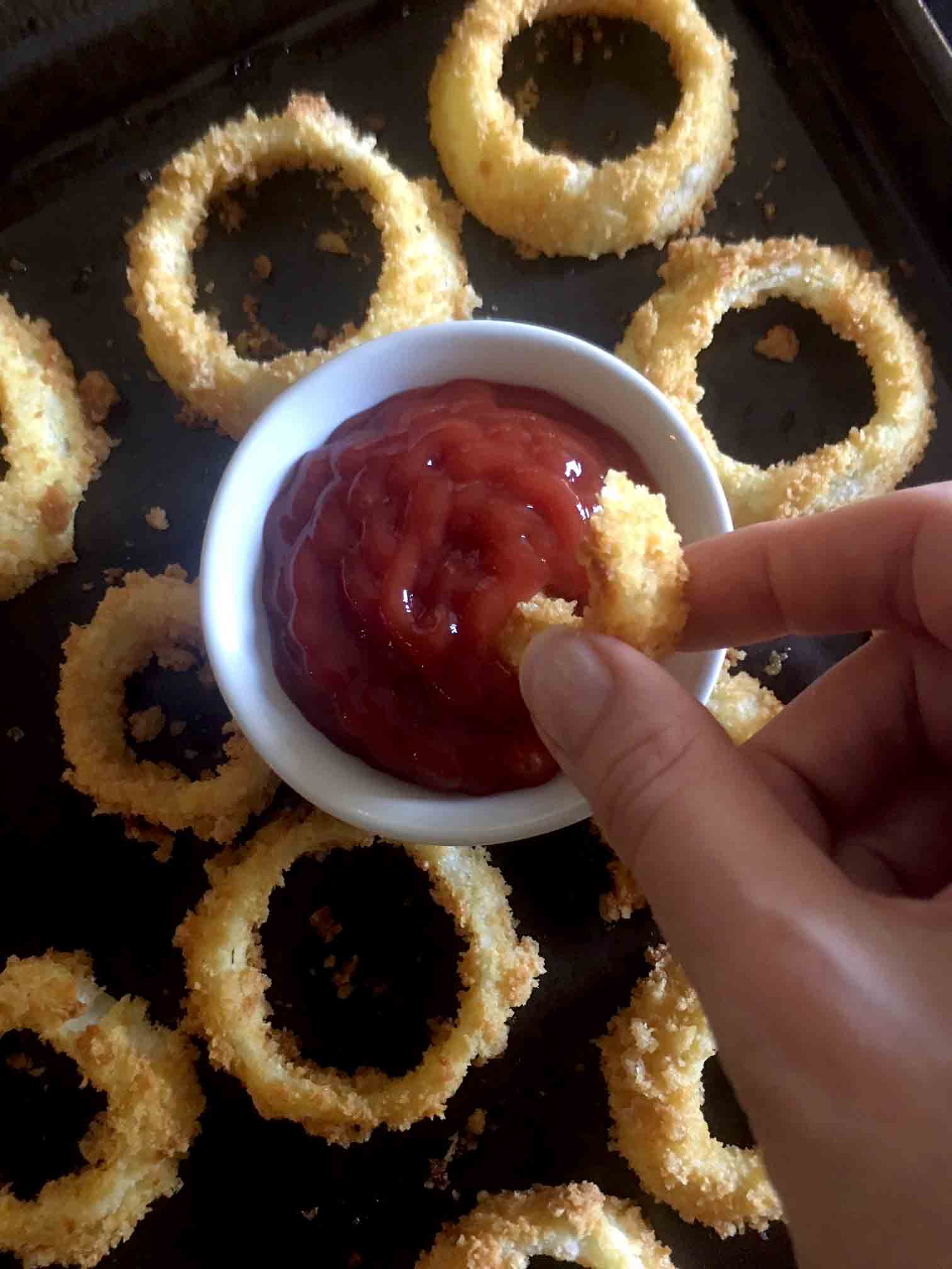 A hand dips a pork rind covered onion ring into a small bowl of sugar free ketchup