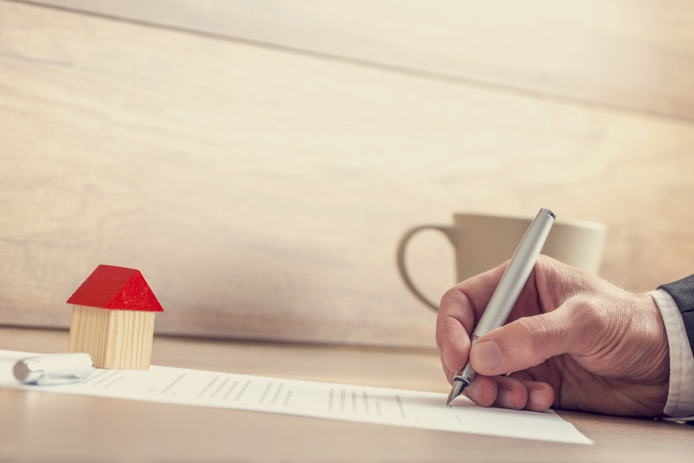Closeup of male hand signing insurance papers, contract of house sale or mortgage documents with fountain pen, wooden toy house sitting on paperwork.
