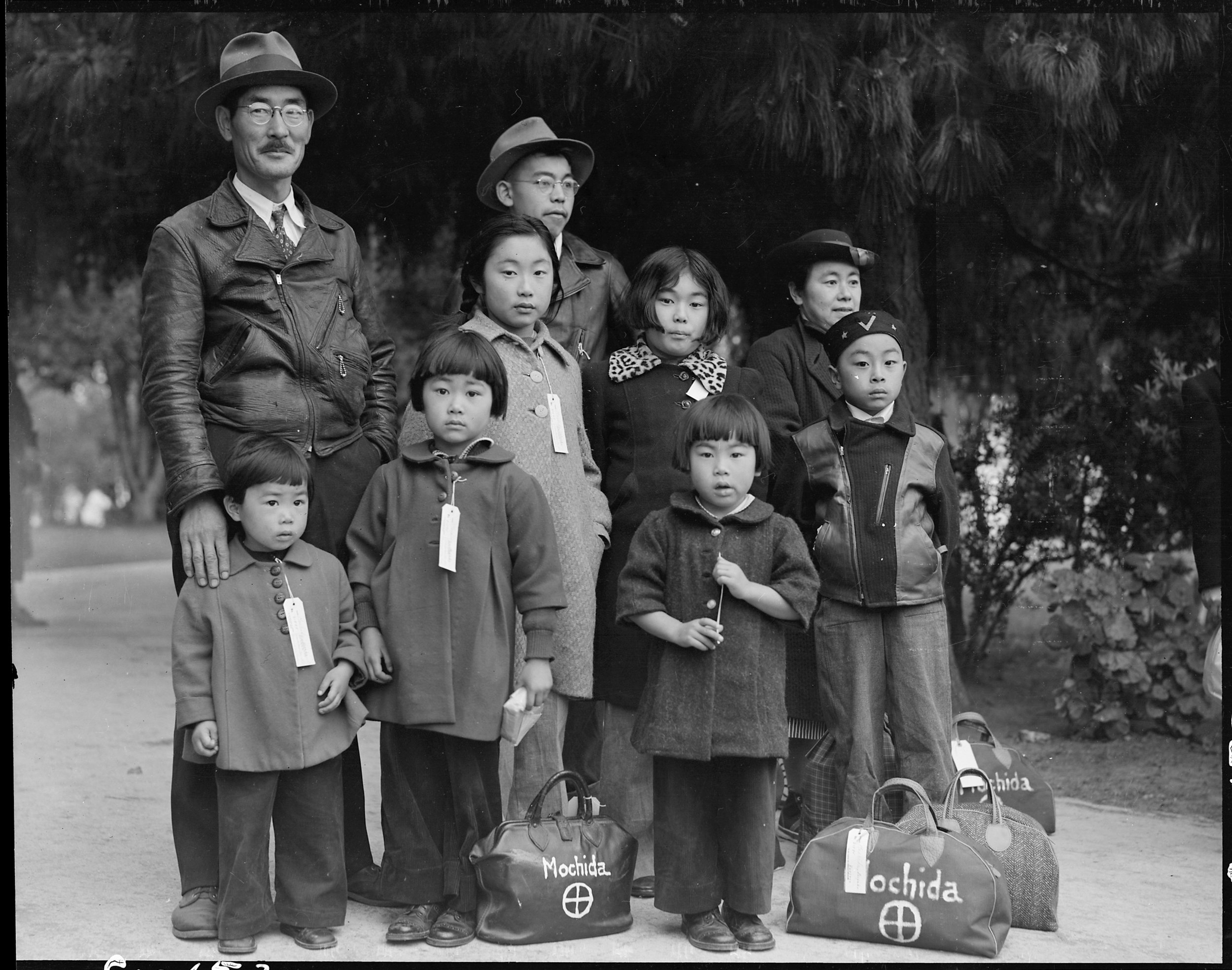 May 8, 1942 — Hayward, California. Members of the Mochida family awaiting evacuation bus. Identification tags are used to aid in keeping the family unit intact during all phases of evacuation. Mochida operated a nursery and five greenhouses on a two-acre site in Eden Township. He raised snapdragons and sweet peas. Evacuees of Japanese ancestry will be housed in War Relocation Authority centers for the duration.