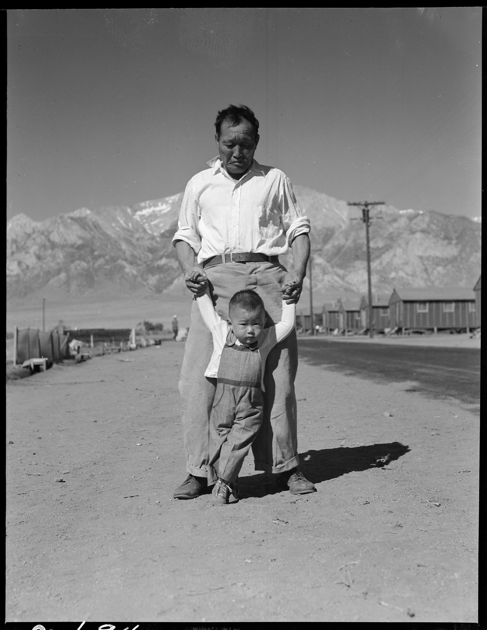 PRINT AVAILABLE July 2, 1942 — Manzanar Relocation Center, Manzanar, California. Grandfather of Japanese ancestry teaching his little grandson to walk at this War Relocation Authority center for evacuees.