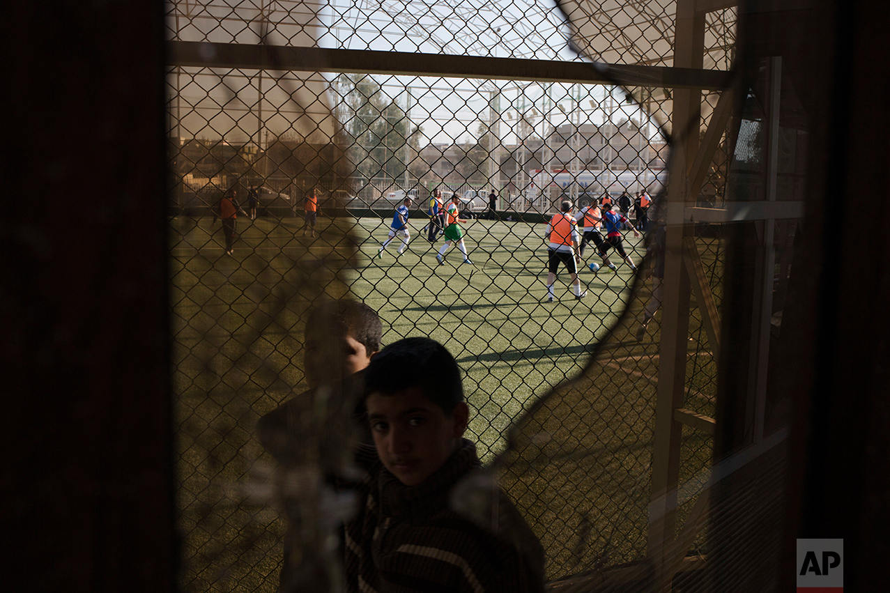A child passes a broken window, damaged by fighting between Iraqi forces and Islamic State militants, while Mosul residents play soccer in the background on Feb. 7, 2017. After months of fighting, Mosul residents can finally practice their favorite game again at the soccer field in the eastern part of the city—and this time without the changes imposed by Islamic State militants. (AP Photo/Bram Janssen)