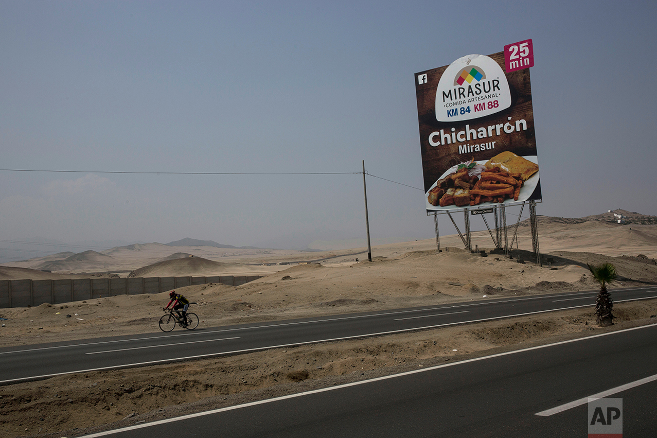 In this May 5, 2017 photo, a cyclist passes a billboard advertising a restaurant along the Pan American Highway on the south side of Lima, Peru. Below the billboards are cannibalized cars, piles of used brick and white crosses marking the places where people died along the highway. (AP Photo/Rodrigo Abd)