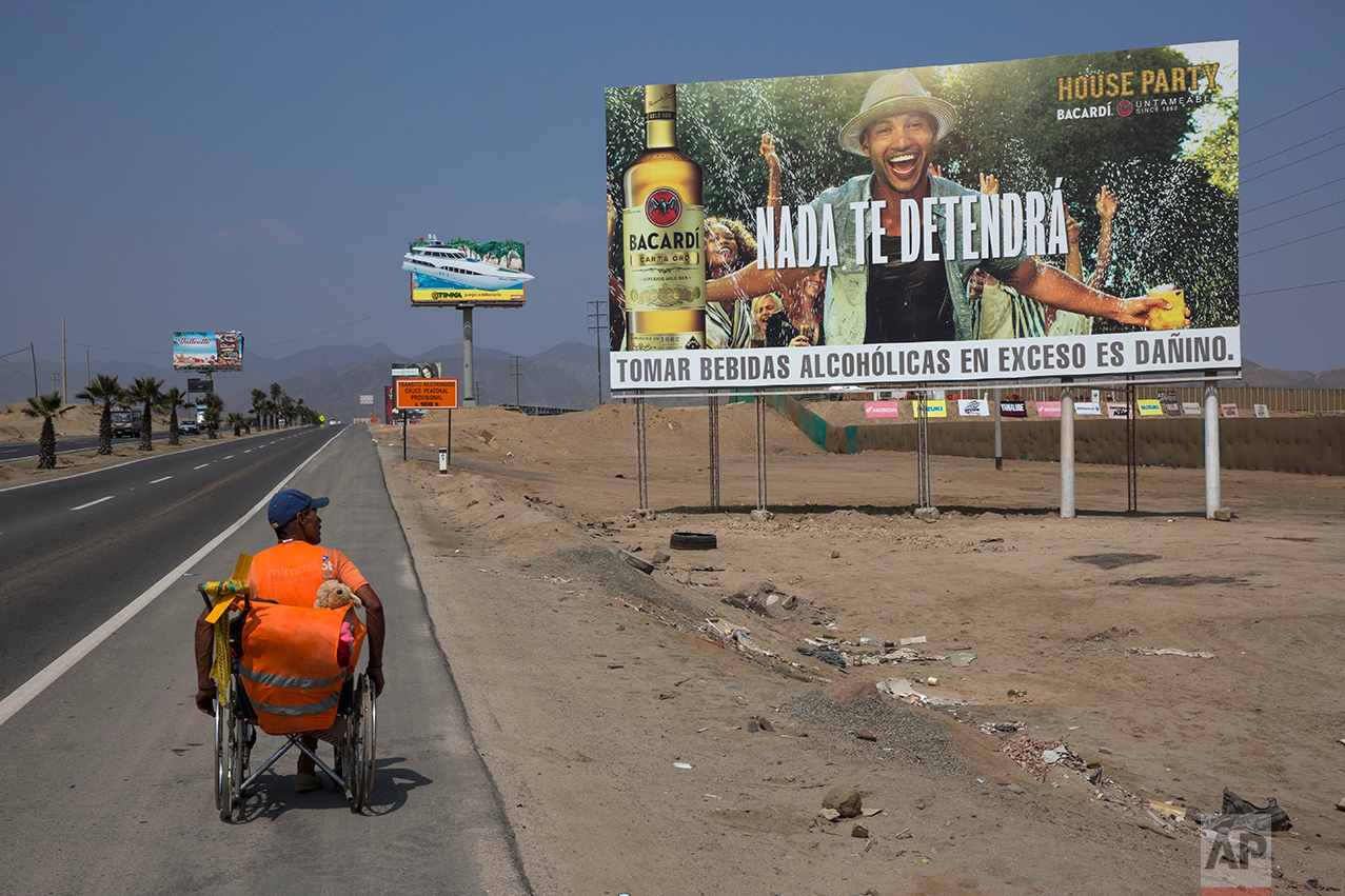 In this May 5, 2017 photo, Jose Suarez, from Cucuta, Colombia, moves his wheelchair, decorated with a stuffed animal, along the Pan American Highway lined with billboards advertising rum, lottery and mixed nuts, on the south side of Lima, Peru. Suarez, 58, said he's on a mission to reach Brazil in his wheelchair, after leaving Cucuta in January. The billboard reads in Spanish "Nothing will stop you." (AP Photo/Rodrigo Abd)