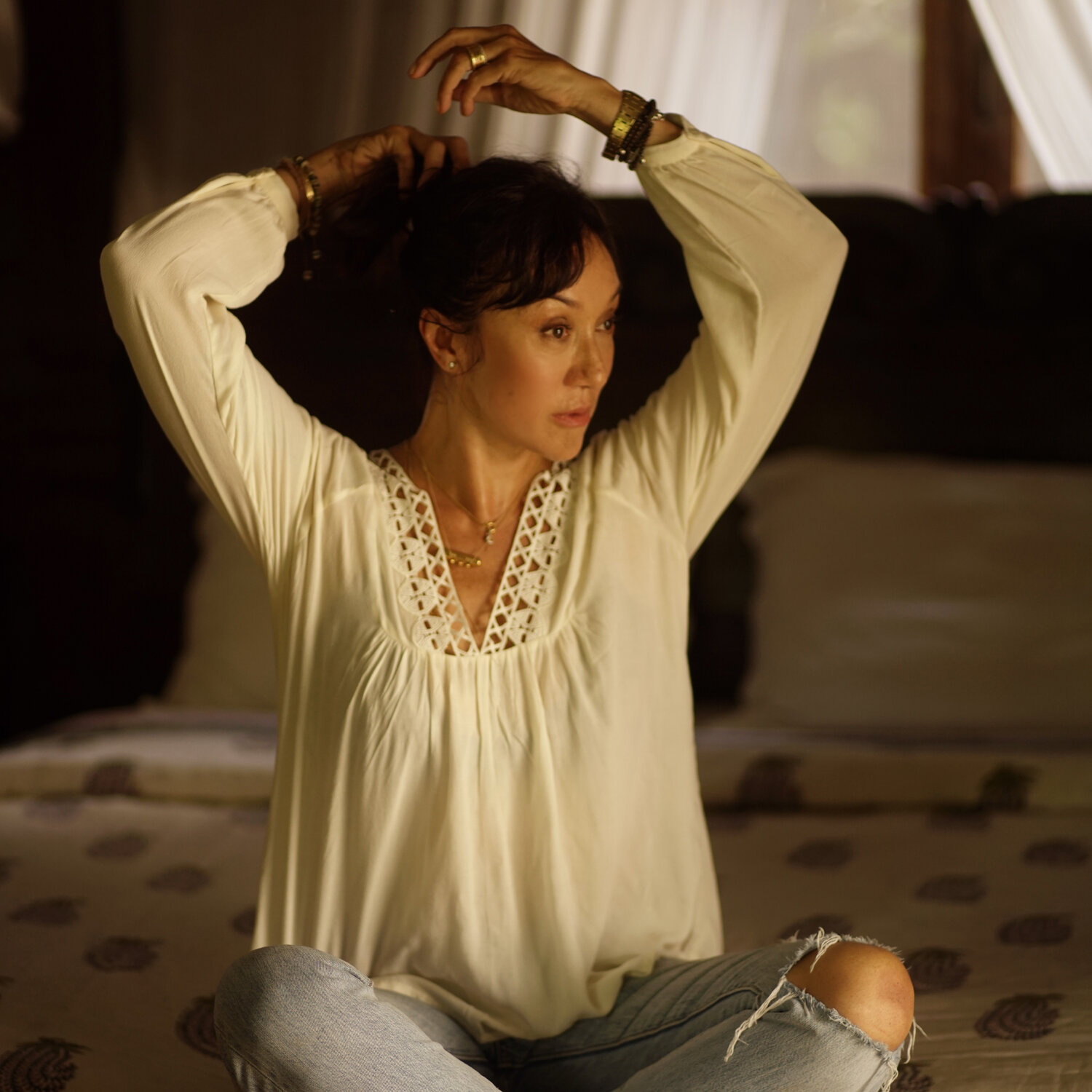 Luiza, the founder of Lulu Yasmine, sits on a daybed wearing light jeans and a delicate white shirt, gracefully putting her hair up. Luiza is a Brazillian-Chinese woman with dark hair.