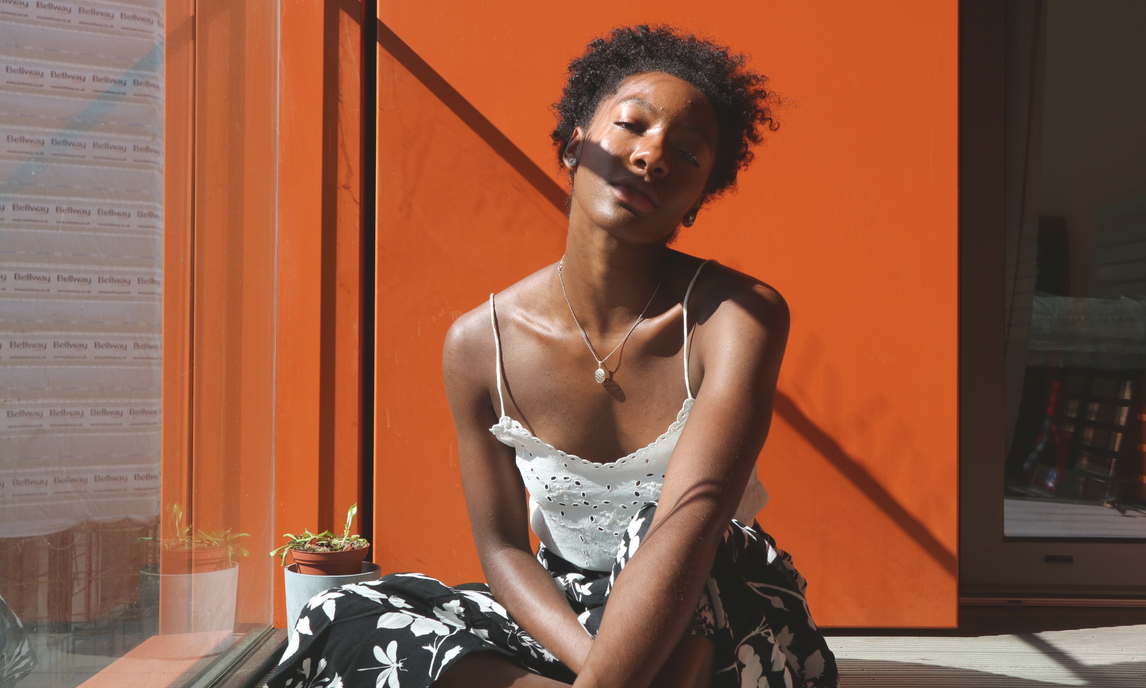Precious is sitting facing the camera wearing a white lacy top and black and white skirt. There is a shadow across her face and an orange wall behind her. Precious is a Black woman with short curly hair.
