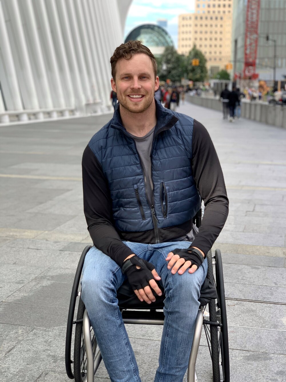 Carson is sitting in his wheelchair on the sidewalk by the Oculus Building in New York, smiling and looking at the camera. He is wearing a blue puffy jacket with black sleeves, a grey shirt, jeans, and fingerless gloves. Carson is a white man with light brown hair.