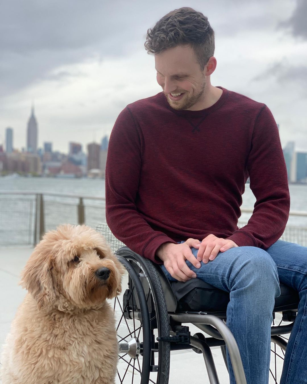 Carson is sitting in his wheelchair by the East River looking down at his dog, Lulu. New York City, including the Empire State building, is vaguely visible in the background. He is wearing a red sweater and light blue jeans. Carson is a white man with light brown hair.