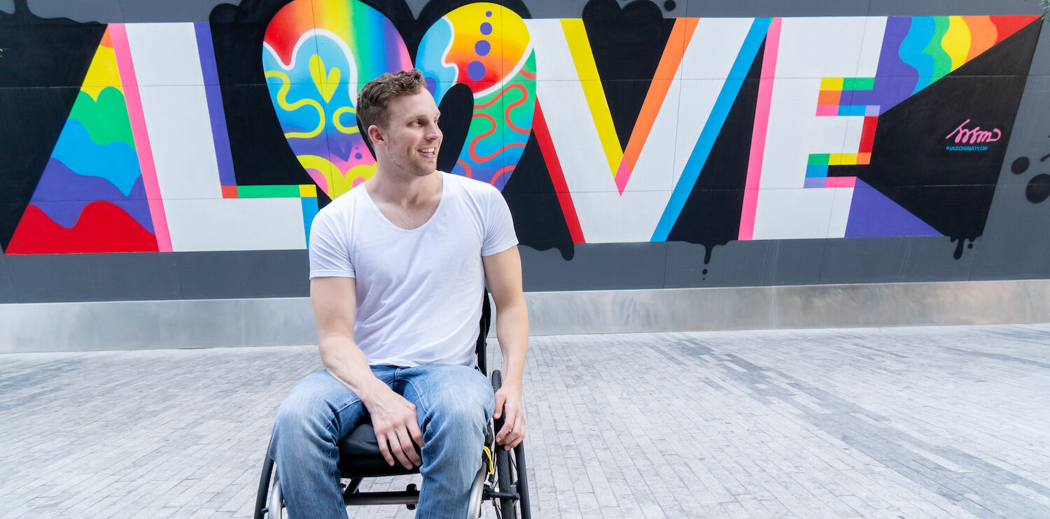 Carson is sitting in his wheelchair in front of a Jason Naylor street mural that says Love in rainbow and white letters on a black background. Fun fact, Jason Naylor is one of my favorite artists. Carson wearing a white shirt and light blue jeans. He is a white man with light brown hair.