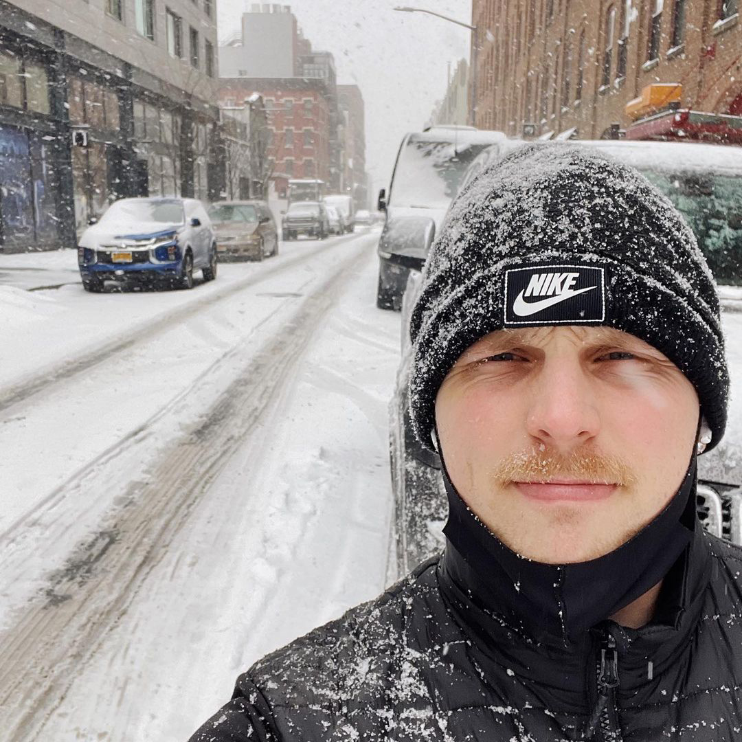 Carson is looking at the camera wearing a black nike hat and black jacket. It's snowing on the New York city street nad he has snow on him. Carson is a white, disabled man with light brown hair.