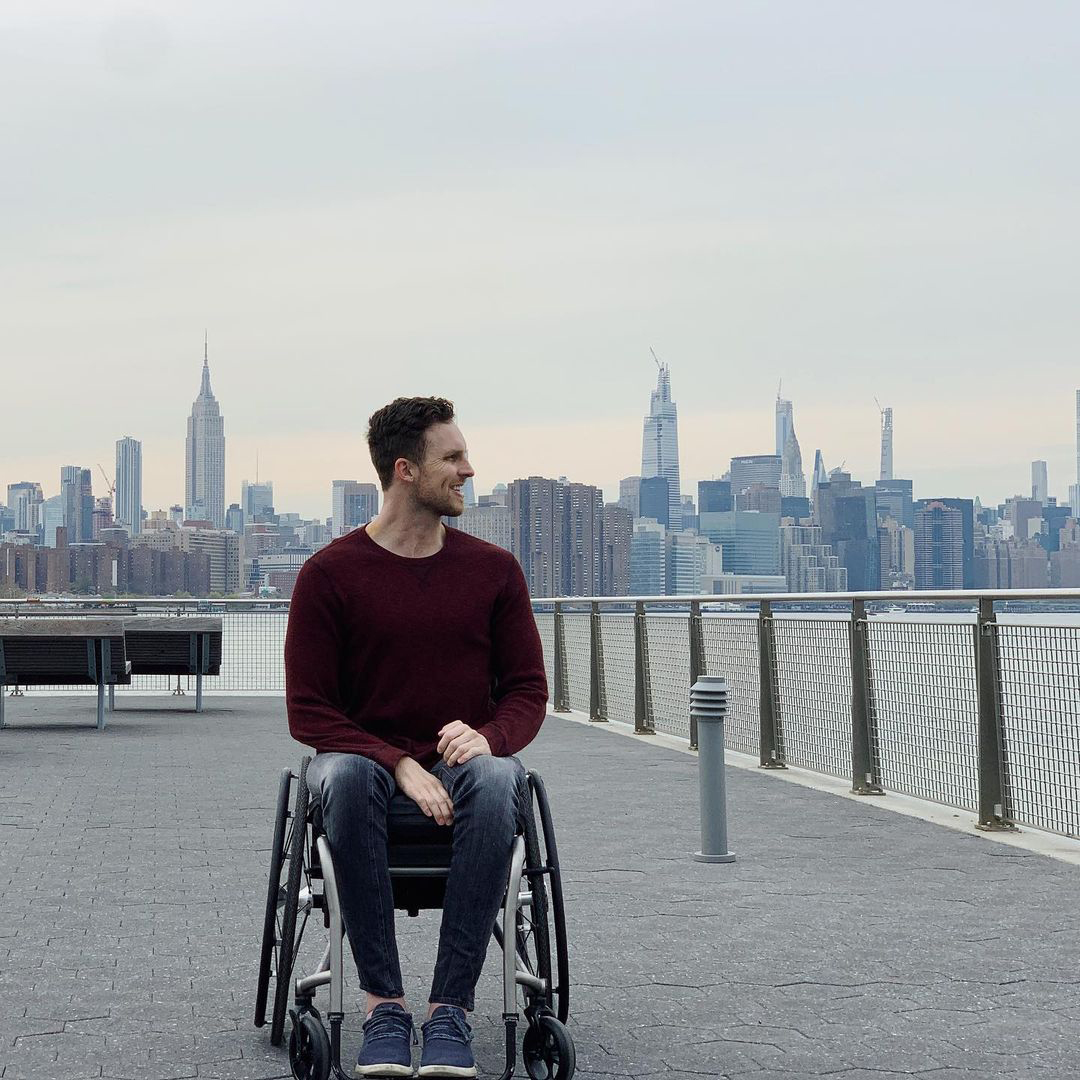 Carson is sitting in his wheelchair looking out at the East River with Manhattan clearly visible in the background. He is wearing a red sweater and blue jeans. Carson is a white man with light brown hair.