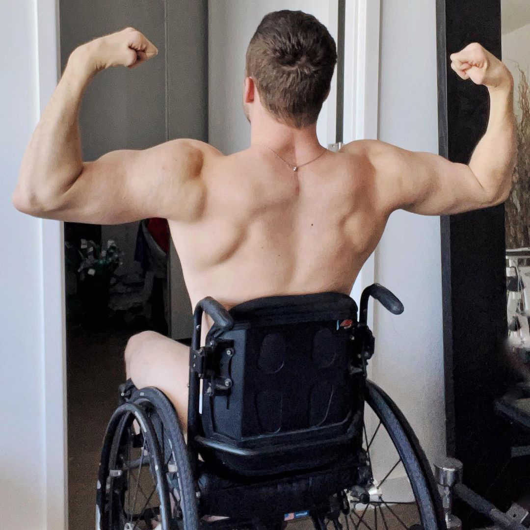 Carson is sitting in his wheelchair facing away from the camera flexing his substantial arm and upper back muscles. He is not visibly wearing antying but a thin necklace. Carson is a white, man with light brown hair.