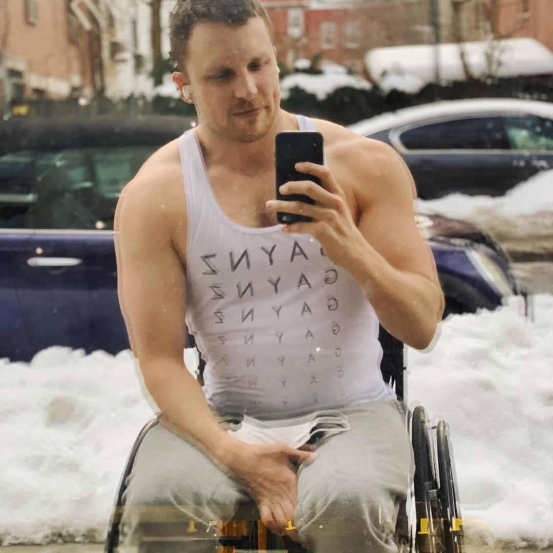 Carson is sitting in his wheelchair taking a selfie with an iPhone in a window on a New York street with snow in the background. He is wearing a white tanktop that says gaynz sevral times down the front, kakhi looking pants, and airpods. There is a white facemask on his lap. Carson is a white, disabled man with light brown hair.
