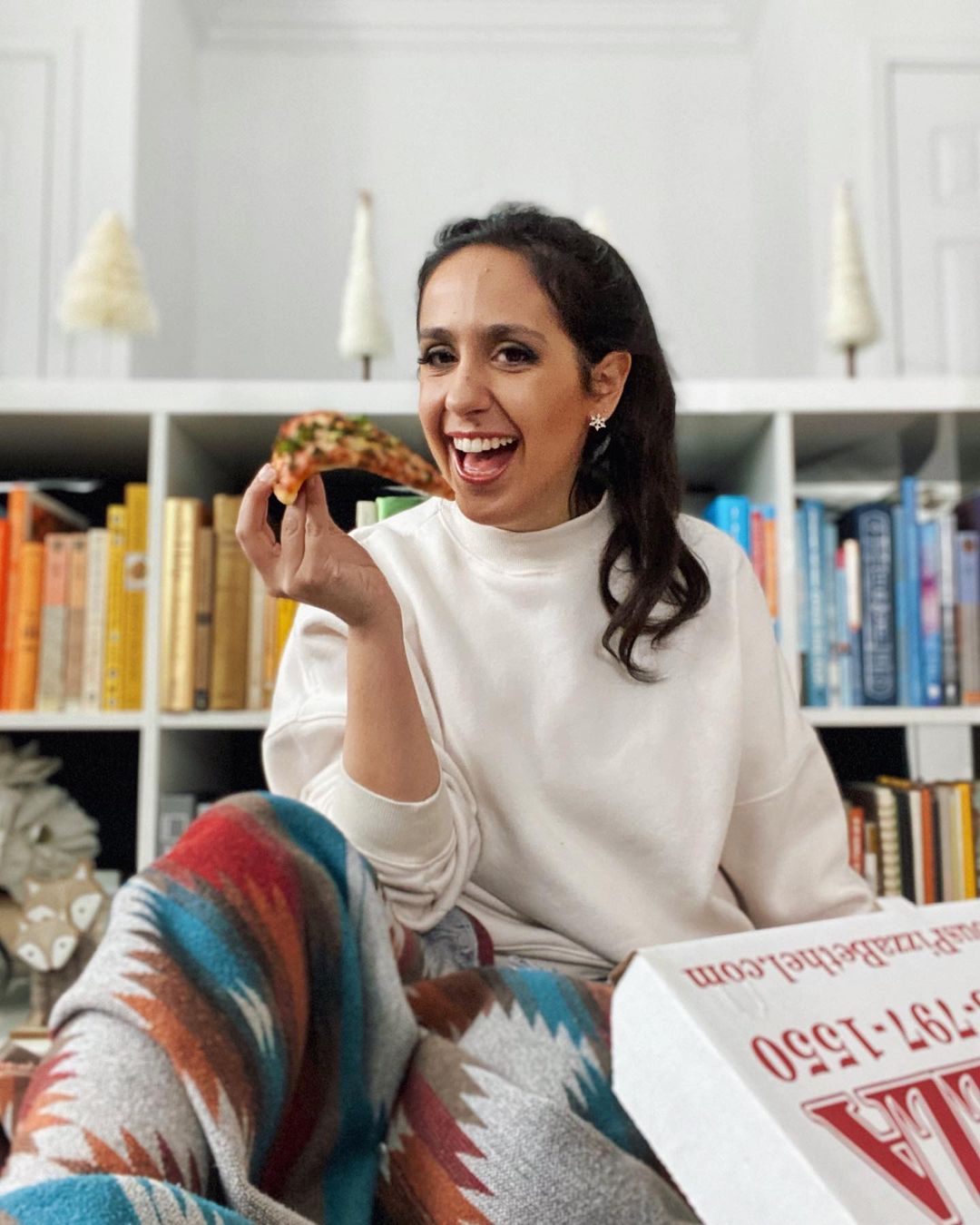 Gabrielle is sitting in front of a bookshelf with books in rainbow order eating a piece of pizza and laughing. Gabrielle is a white woman with long brown hair in a ponytail, wearing a white sweatshirt and a blanket from Mini Tipi, an awesome indigenous-owned company I highly recommend you check out.
