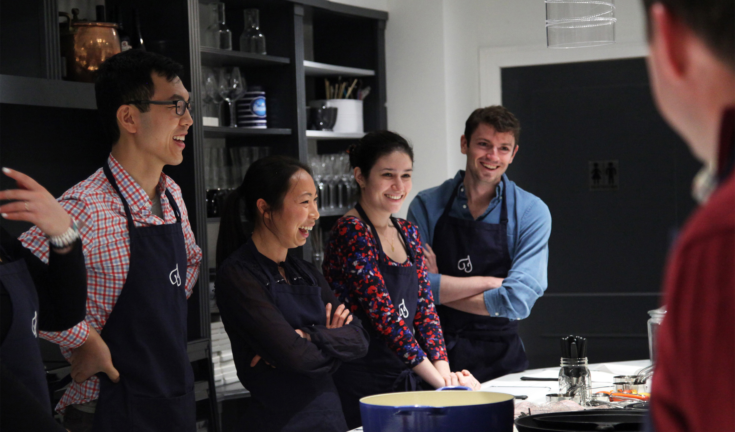 Four people in their twenties are standing and laughing at something. They are all wearing navy blue aprons with the Fig Cooking School logo on them. This is the cover picture on the Fig Cooking School Website.