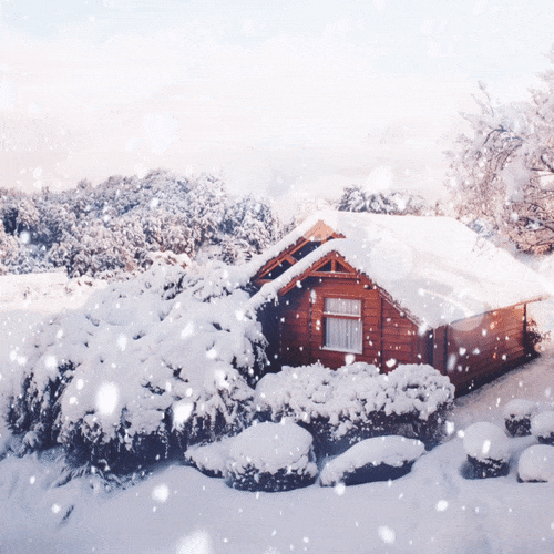 A GIF of a ski chalet with constantnly falling snow over it, and a bright lens flare over the top. There is snow on the roof and snowy trees in the background.