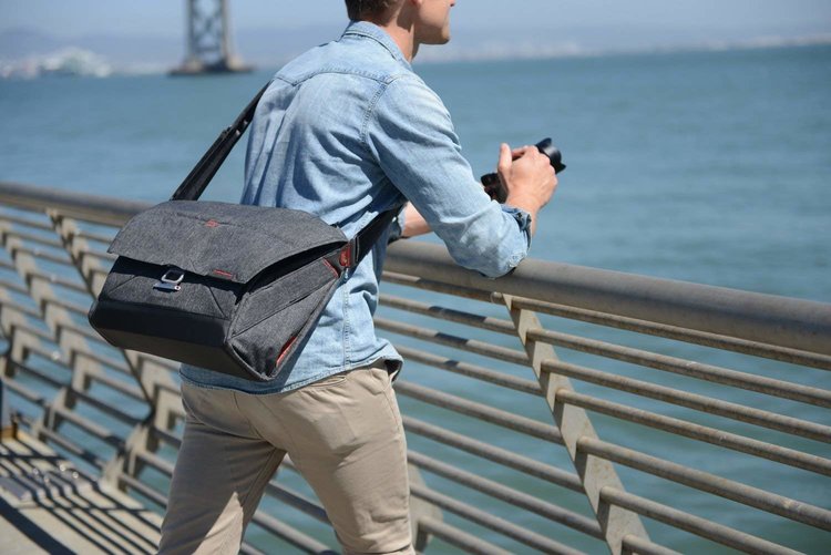 Image of a man holding a camera next to the ocean, with a 15" Everyday Messenger camera bag slung onto his back.