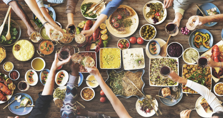 Overhead shot of hands reaching for various dishes of food on a table, and also cheersing wine glasses