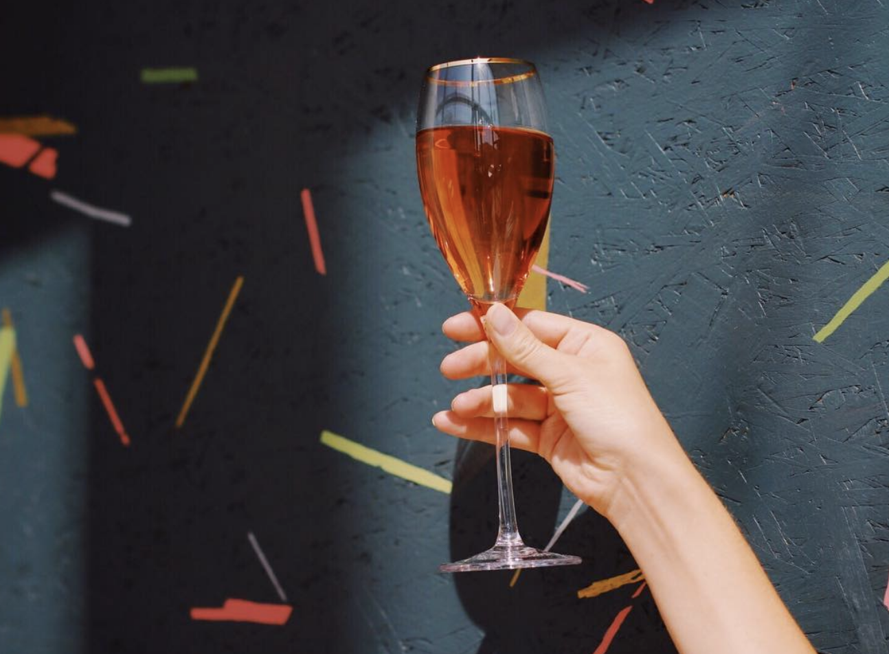 Woman's hand with painted nails holding up a champagne flute of rosé champagne in front of a dark painted wall