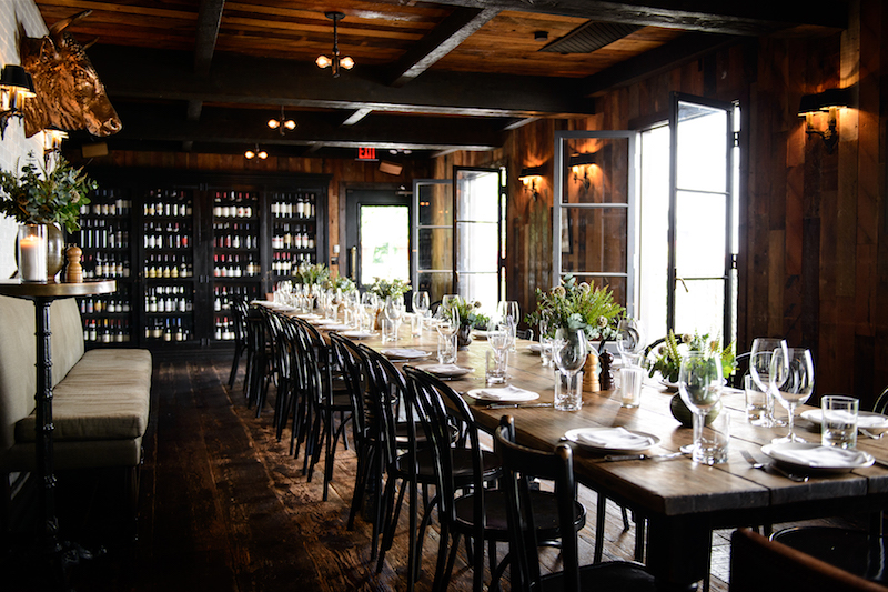 Interior of a restaurant with a long table with chairs, and a wall of wine bottles