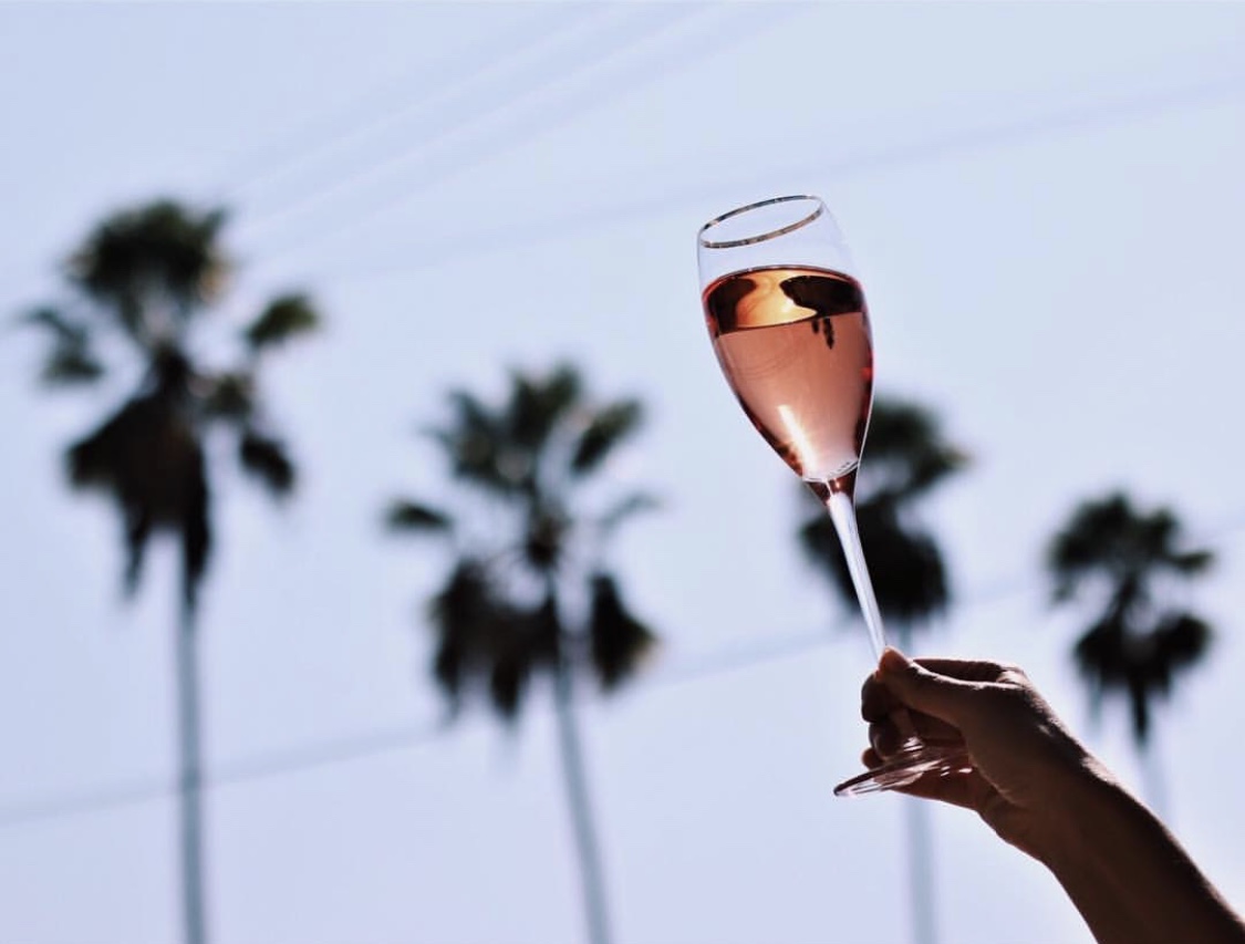 Woman's hand holding up a wine glass of rosé wine in front of a sky backdrop with 4 palm trees in silhouette