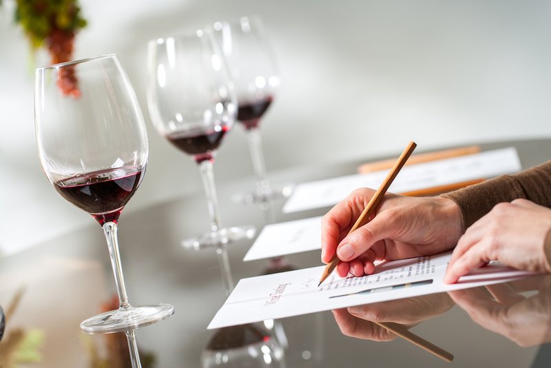 three glasses of red wine in a row in front of hands holding a pencil and answering questions on a sheet of paper