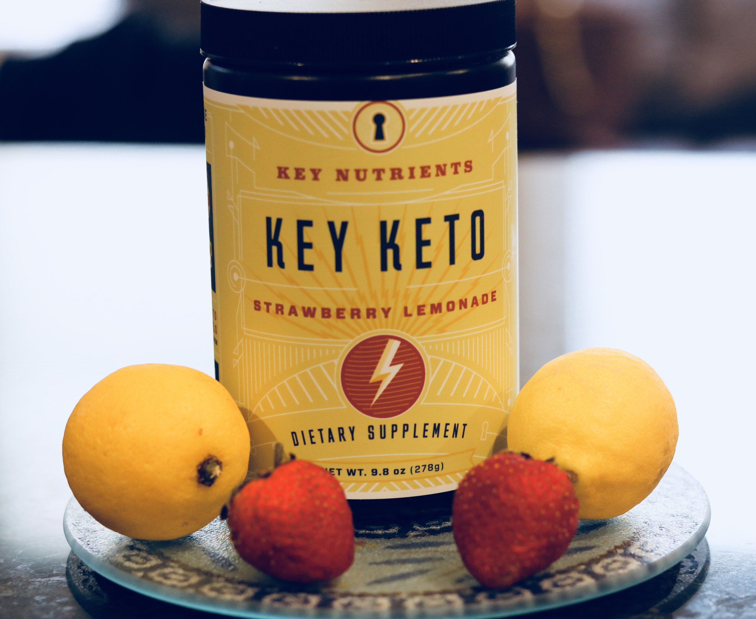 A container of Key Keto\'s Strawberry Lemonade power, along side two lemons and two strawberries