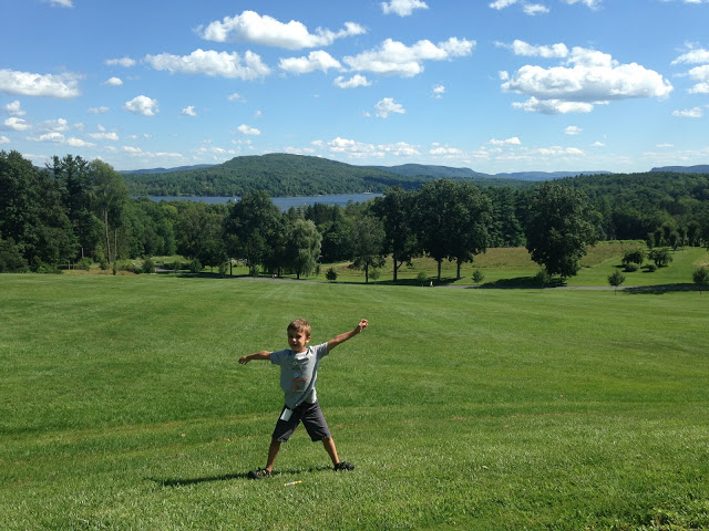 5 year old Eli standing with his feet wide and his arms open in a green field with trees and hills in the distance