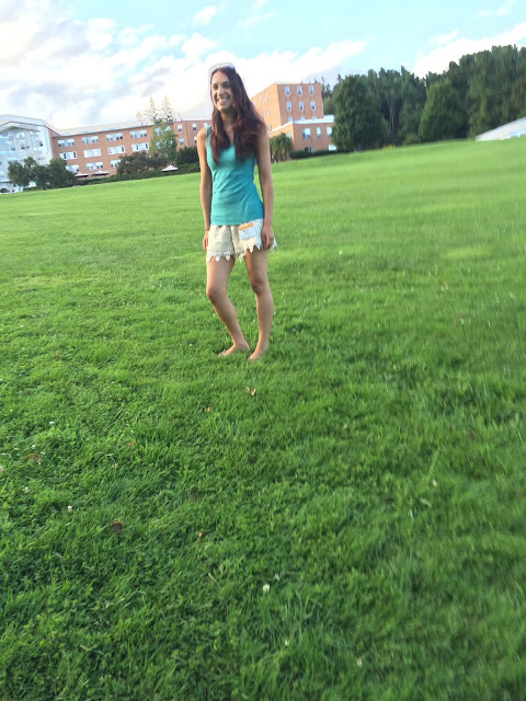 Marisa smiling standing in the grass wearing a teal tank top and white shorts