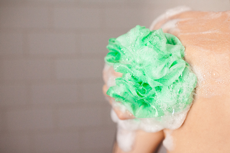 Model washing shoulder using green loofah in shower.