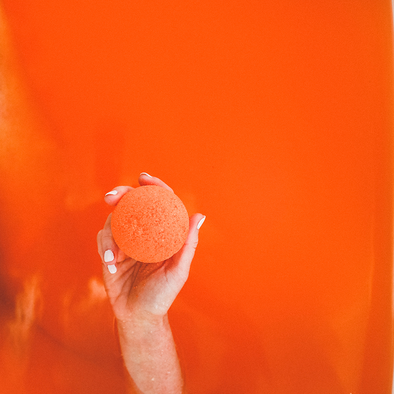 Model holds orange Citrus CBD bath bomb out of orange bath tub water. 