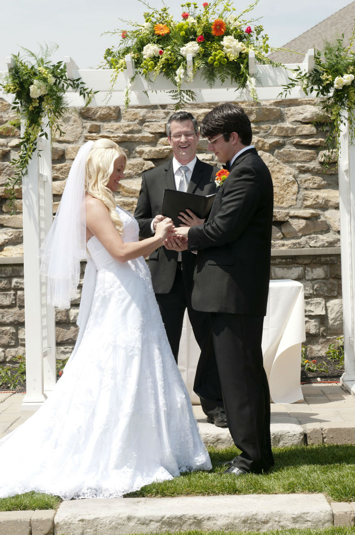 Wedding officiant Damian King smiles as Teresa places ring on Ben's hand