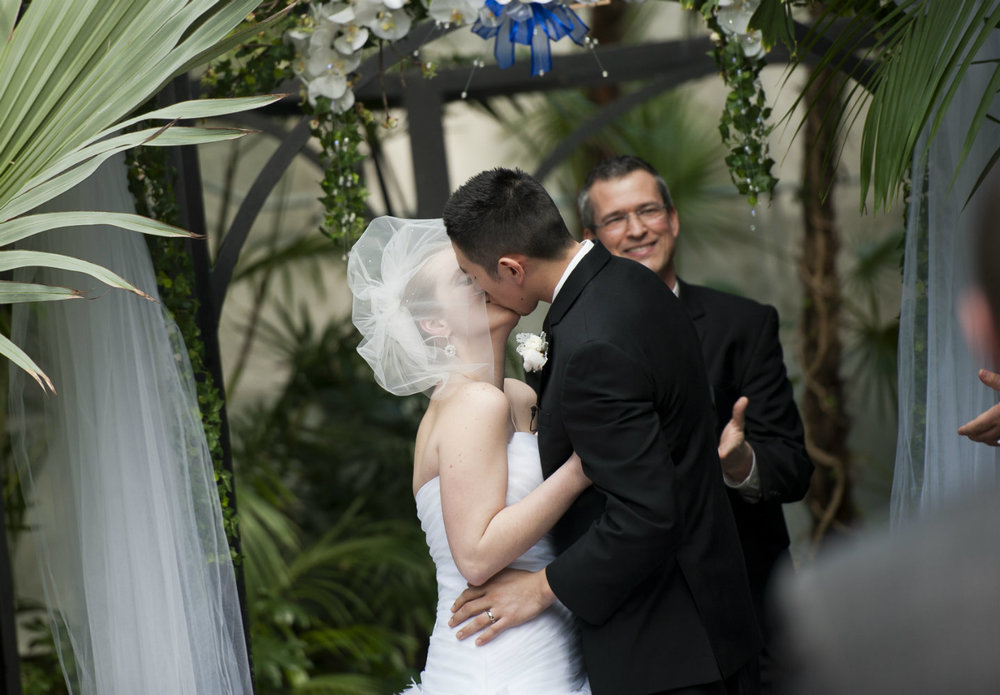 Wedding officiant Damian King smiles as bride & groom kiss in Palm House