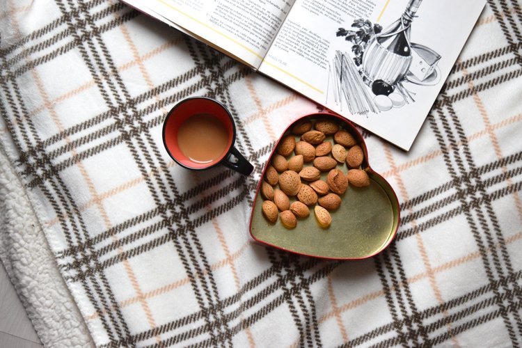 Almonds In A Heart-Shaped Container, A Coffee Mug, And A Cookbook On A Quilt.